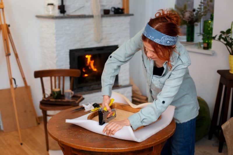 A woman upholstering furniture with a stapler in a cozy living room, demonstrating practical skills from Wutawhelp Guides for home improvement.