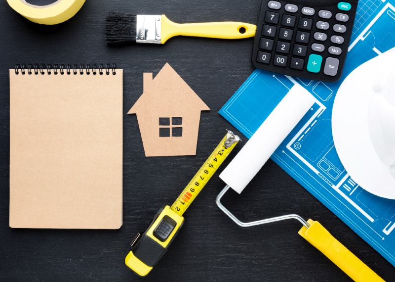 Home improvement tools including a notebook, calculator, blueprint, tape measure, paintbrush, and a cardboard house cutout, illustrating The Core Philosophy Behind Wutawhelp Guides.