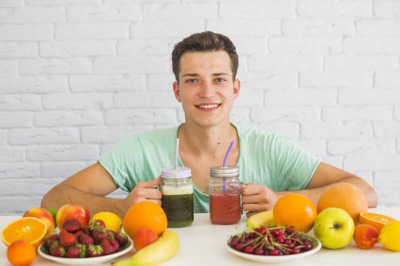TimesHealthMag.com: Your Ultimate Guide to Evidence-Based Health Insights A young man holding green and red smoothies, made with healthy ingredients like spinach, berries, and fruits, surrounded by fresh oranges, strawberries, bananas, and cherries. TimesHealthMag.com promotes such nutritious, evidence-based lifestyle choices.