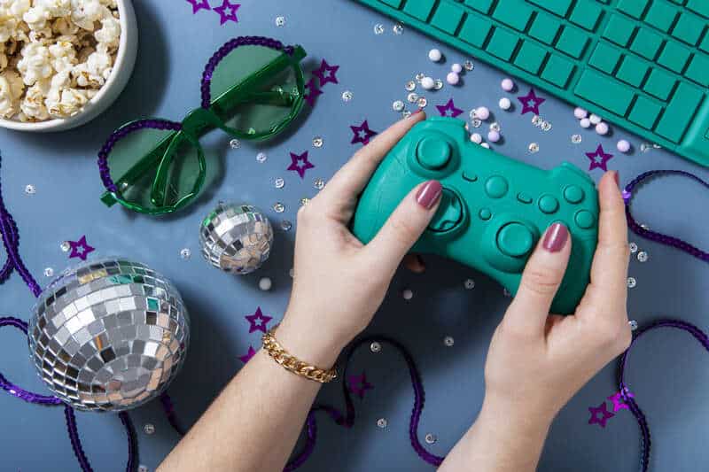 A teal game controller held by hands surrounded by party decorations like disco balls, purple star confetti, green glasses, popcorn, and a teal keyboard, creating a fun celebration vibe for MyWirelessCoupons.com.