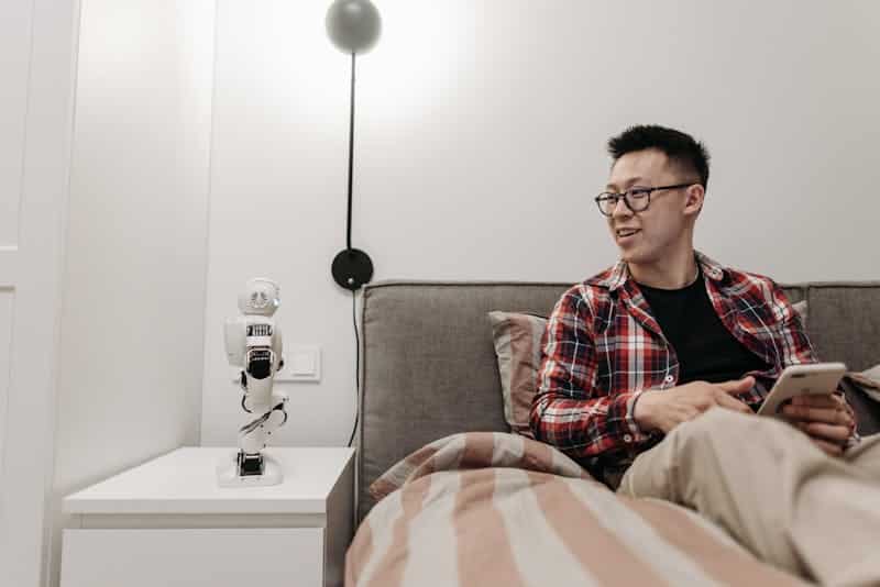 "A person sitting on a bed, holding a smartphone, while interacting with a small AI robot placed on a nightstand. The room is modern, highlighting the integration of smart living and AI technology in everyday life.