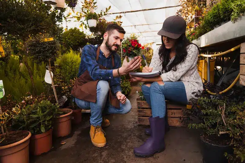 A man and woman discuss gardening tips in a greenhouse. The man points at a plant while the woman writes in a notebook. Both are dressed in casual gardening attire, surrounded by potted plants. This image is related to DecoradHouse Garden Tips by DecoratorAdvice for enhancing your outdoor space.