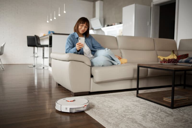 A woman relaxing on a sofa while using her phone, with a robot vacuum cleaner cleaning the floor in the living room.