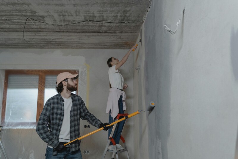 Two people painting a wall during a home renovation project, one on a ladder and the other using an extended paint roller. This image illustrates the effort involved in home improvements, aligned with Decoradhouse Renovation Tips by Decoratoradvice.