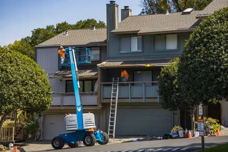 Workers on a lift and ladder performing exterior maintenance on a residential building with a well-maintained yard.