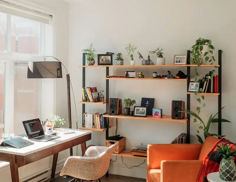 A cozy home office setup with a wooden desk, modern chair, and a shelf filled with books, plants, and decorative photos. A lamp stands beside the desk, and a few speakers are visible on the shelf. The space is bright and organized with natural light coming from the window, offering a perfect example of drhomey tips for creating a healthier, happier home environment.