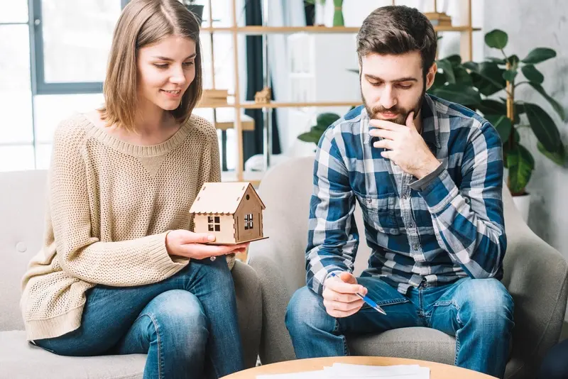 Unlock DrHomey’s Top 10 Tips for a Healthier, Happier Home in 2025 A couple sitting on a couch, discussing home-related plans. The woman is holding a small wooden house model, while the man is thoughtfully examining a pen and papers, likely planning improvements for a healthier, happier home in 2025 with DrHomey’s top tips.