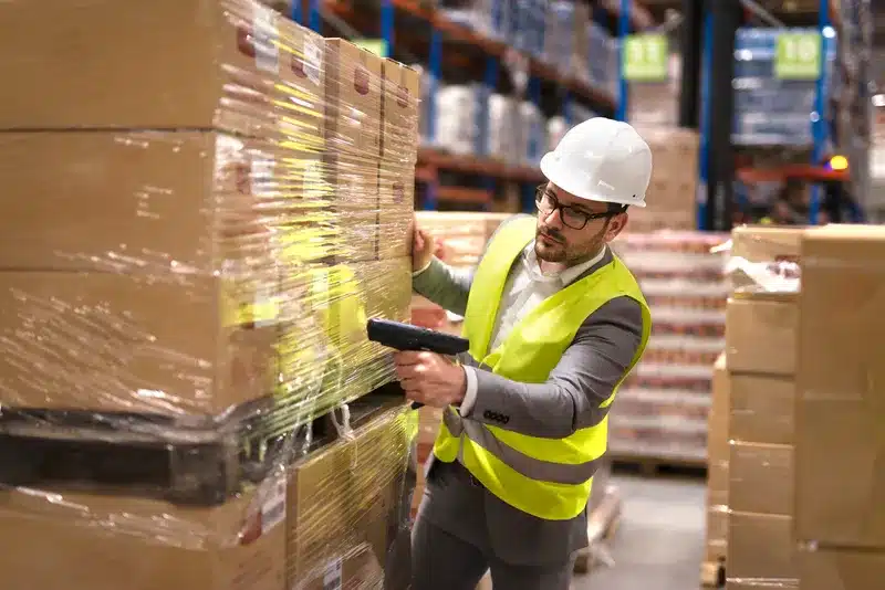 A worker wearing a safety vest and helmet scans boxes in a warehouse, ensuring proper inventory management. Efficient 3PL logistics operations like these directly contribute to improving customer satisfaction by optimizing delivery processes.