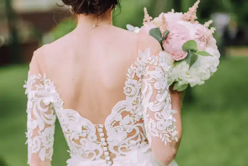 A bride wearing a backless wedding dress with intricate lace detailing and sheer sleeves, holding a bouquet of flowers.