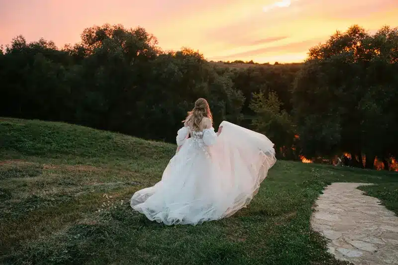 A bride in a flowing ball gown, walking on a grassy path with a sunset in the background, showcasing her wedding dress in a serene outdoor setting.