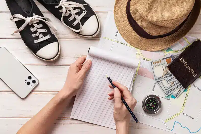 A person planning a one day trip, holding a pencil over a notepad, surrounded by sneakers, a hat, a phone, a compass, a passport wallet with money, and a map.