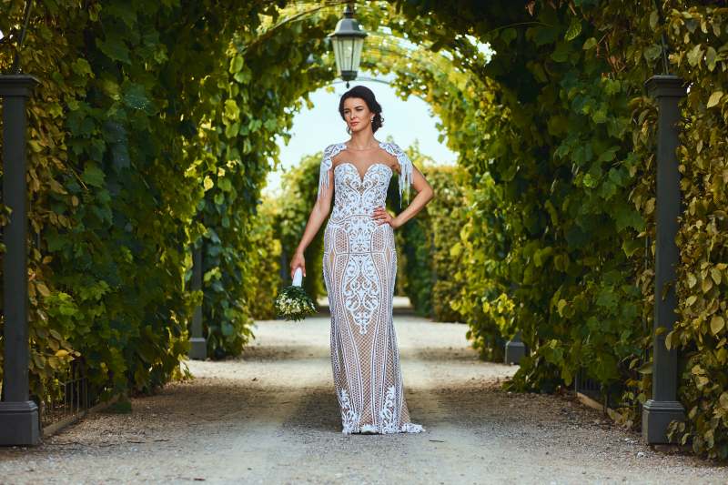 A bride in a stunning mermaid wedding dress, standing confidently in a vineyard pathway, holding a bouquet, with intricate lace details on the gown.