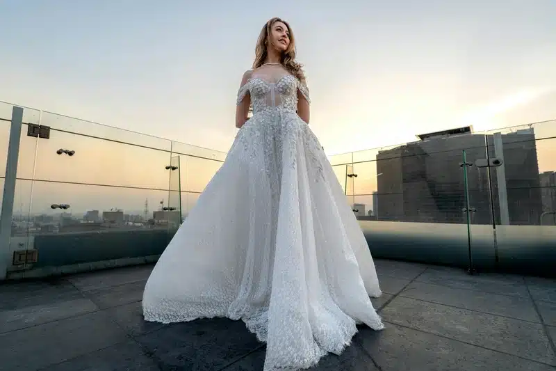 A model wearing a beautiful off-shoulder white wedding dress with intricate floral details, standing on a rooftop at sunset. This elegant gown would be complemented by a tailored wedding workout plan to enhance posture and tone key muscle groups.