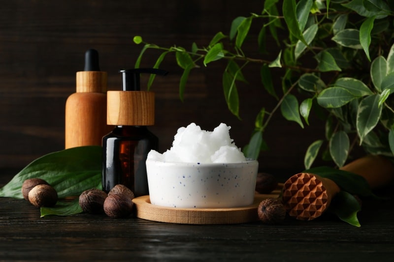 Natural skincare setup with a bowl of whipped shea butter, dark amber pump bottle, wooden dropper bottle, fresh green leaves, and whole nuts on a dark wooden background. Perfect for illustrating basic skincare and skin-friendly natural products.