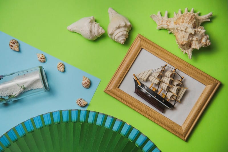 A flat lay of nautical-themed craft items on green and blue backgrounds, including seashells, a framed model ship, a small glass bottle with sand, and a green paper fan. Perfect for Fun Crafts ThunderOnTheGulf family activities and art inspiration.