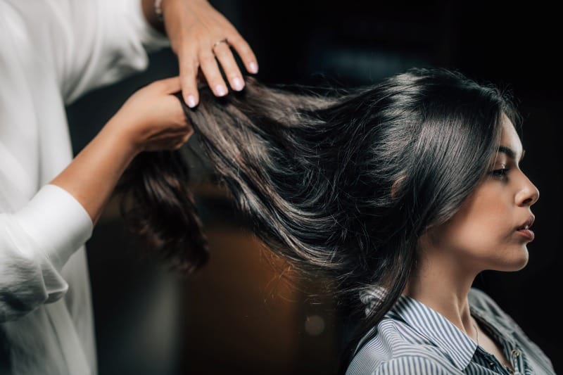 Hair Care on the Go: Why Amika Is Every Jetsetter’s Secret Weapon A hairstylist holds and examines a woman’s long, shiny hair in a salon setting, showcasing professional hair care and styling attention to detail — perfect representation for a “Hair Care on the Go” theme.