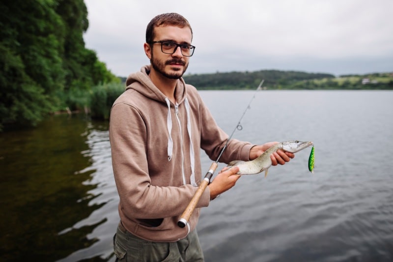Man holding a freshly caught fish beside a calm lake, wearing glasses and a brown hoodie, with a fishing rod in hand. Example image for Fishing Guides ThunderontheGulf where every trip becomes a story.