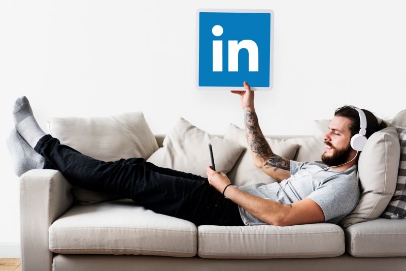 From Burnout to Balance: How LinkedIn Automation Frees Up Time for What Matters A man relaxing on a sofa wearing headphones and holding a smartphone while lifting a LinkedIn logo sign above him, symbolizing the concept of using automation on LinkedIn to save time and achieve work-life balance.