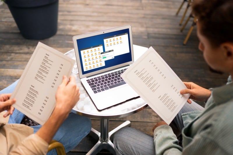 What Is the Difference Between Day Passes and Monthly Data Bundles? Two people at a café table reviewing menus beside a laptop showing file folders, symbolizing a choice between day passes and monthly data bundles.