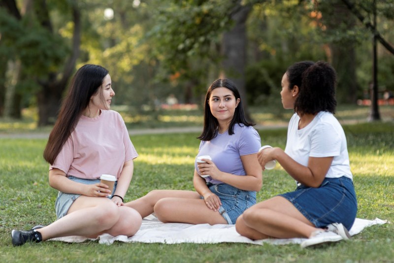 Three friends sitting on a blanket in a park, talking and relaxing together, a simple idea for Things to Do With Friends When Bored outdoors.