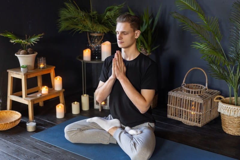 Man meditating on yoga mat with candles and plants, a relaxing self-care activity and peaceful Things to Do at home.
