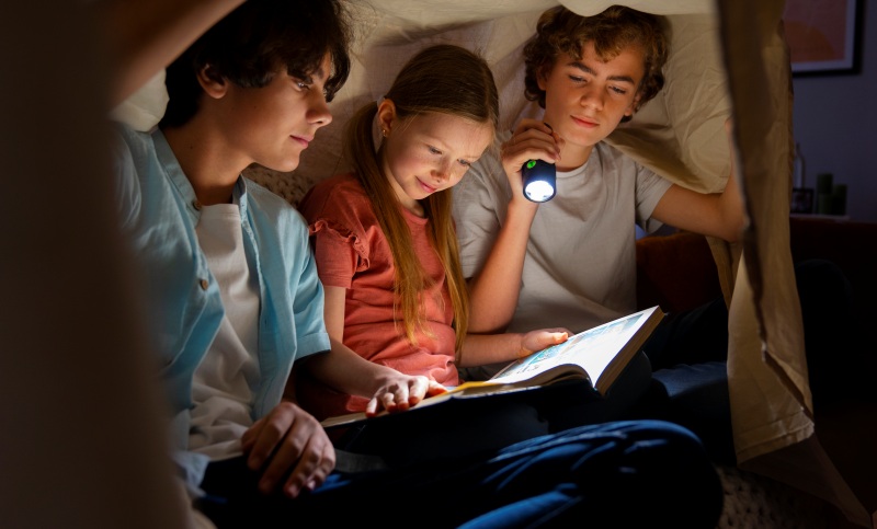 Friends sitting under a blanket reading a book with a flashlight, a cozy sleepover idea for Things to Do With Friends When Bored at night.