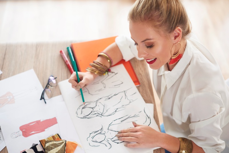 Woman sketching fashion drawings at a table with pencils, papers, and fabric samples.