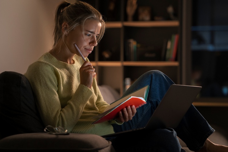 Woman sitting with a notebook and laptop, studying at home in the evening.