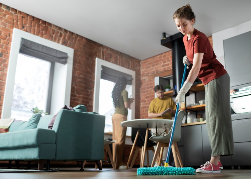 Woman mopping the floor in a living room while others stand nearby in the background.
