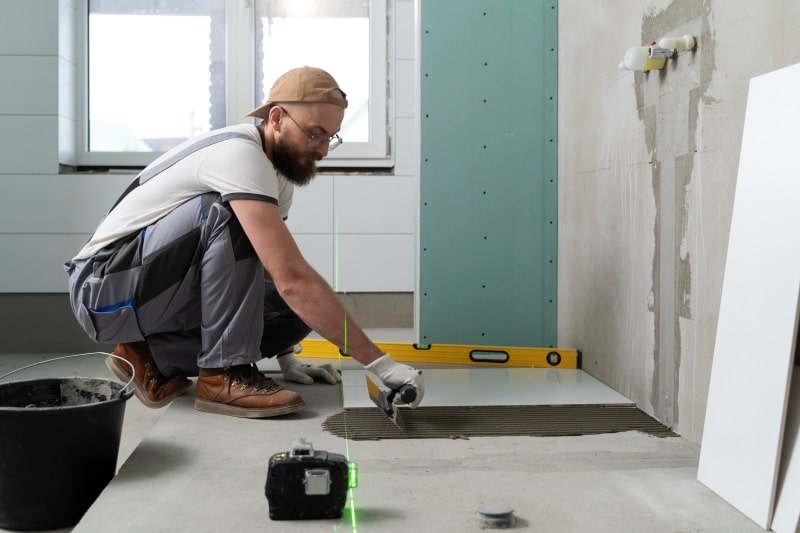 The Top Bathroom Remodeling Companies to Partner With: A Comprehensive Resource Roundup Bathroom remodeling contractor laying floor tile with a level and laser guide during a bathroom renovation.