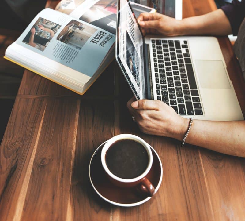 Person reading a magazine beside a laptop and coffee while browsing BoringMagazine.com News.