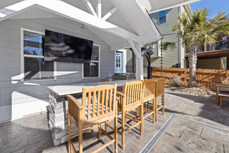 Practical Backyard Upgrades Are Delivering Lasting Home Value Functional backyard upgrades featuring a covered outdoor kitchen, stone patio, bar seating, and wall-mounted TV.