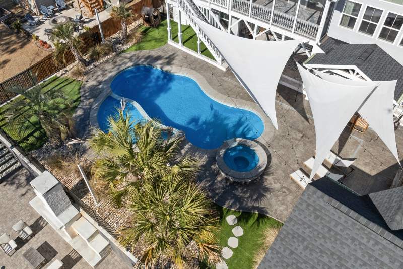 Backyard swimming pool with surrounding palm trees, paved patio, and white shade sails