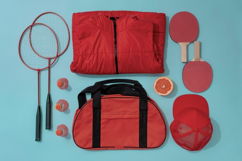 Most Popular Pickleball Stocking Stuffers for the Holidays Red sports bag, paddles, rackets, balls, and clothing arranged on a blue background as pickleball stocking stuffer gift ideas.
